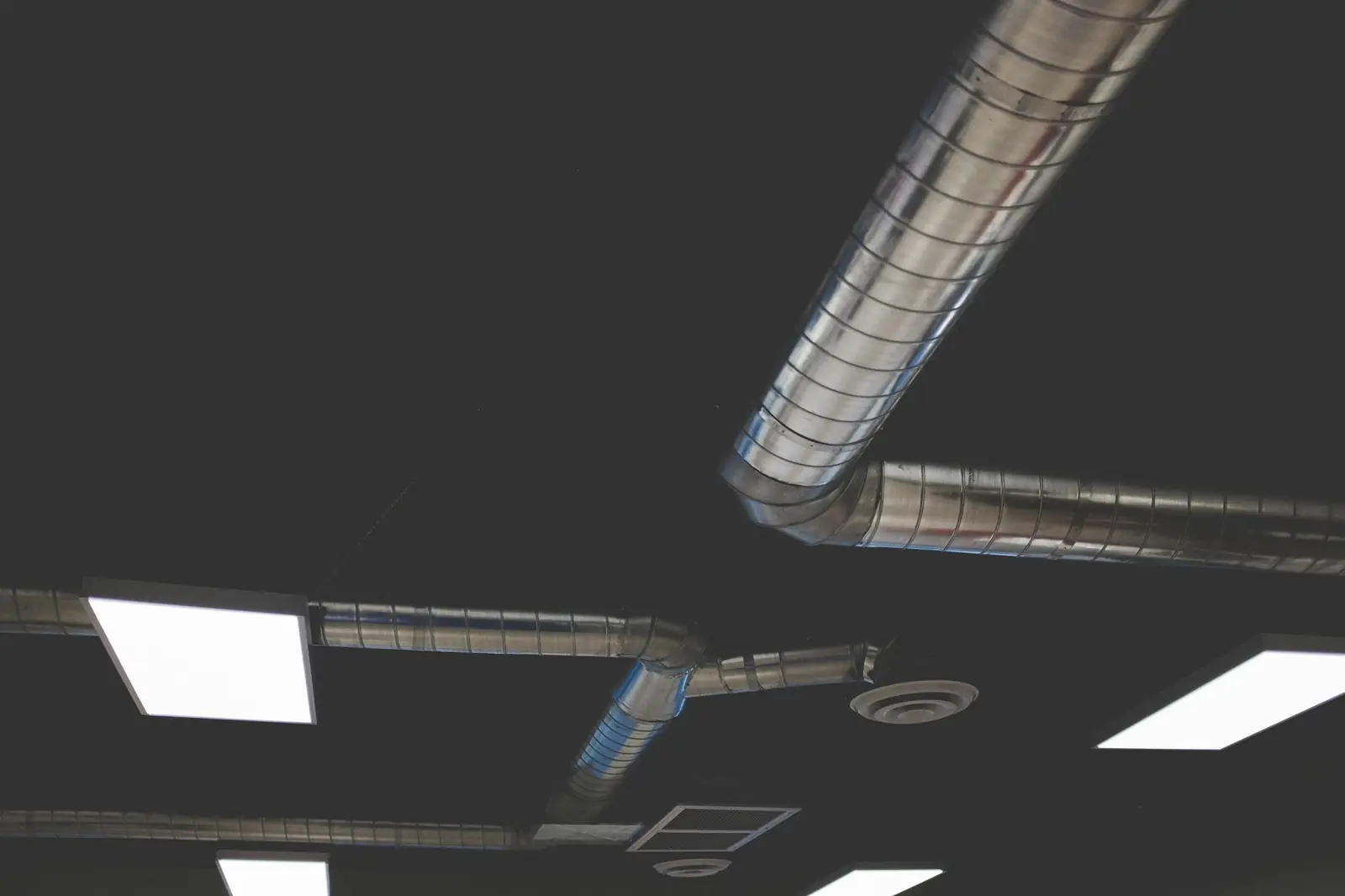 Spiralfalsade ventilationskanaler i blank plåt och takdon i ett mörkt innertak med LED-paneler